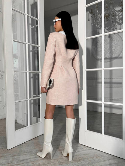 Woman in a light pink coat and white knee-high boots standing in a room with large windows.