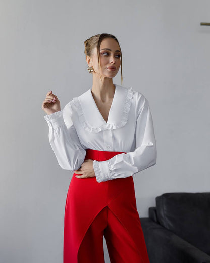 Woman wearing a white blouse with ruffled collar and red pants against a plain background