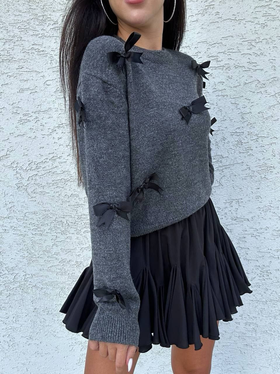 Person wearing a gray sweater with black ribbons and a black skirt against a light gray background