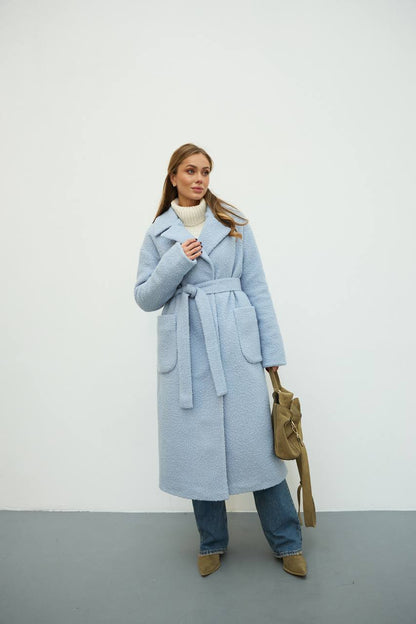 Woman wearing a light blue coat with a belt, standing against a white background.