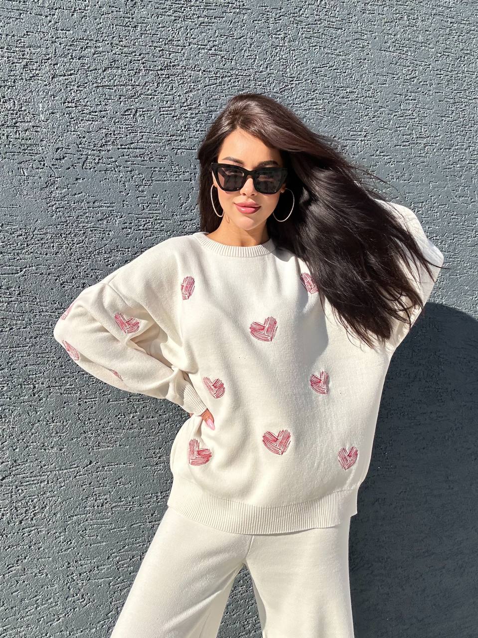 Woman wearing a white sweater with pink heart patterns against a gray wall.