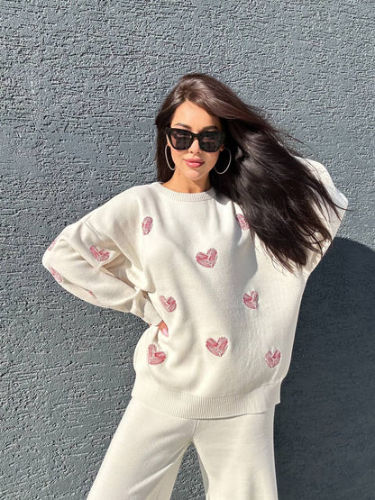 Woman wearing a white sweater with pink heart patterns against a gray wall.
