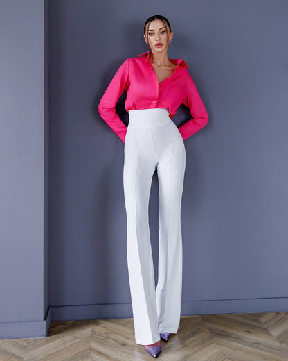 Woman wearing a bright pink blouse and white pants against a gray wall.