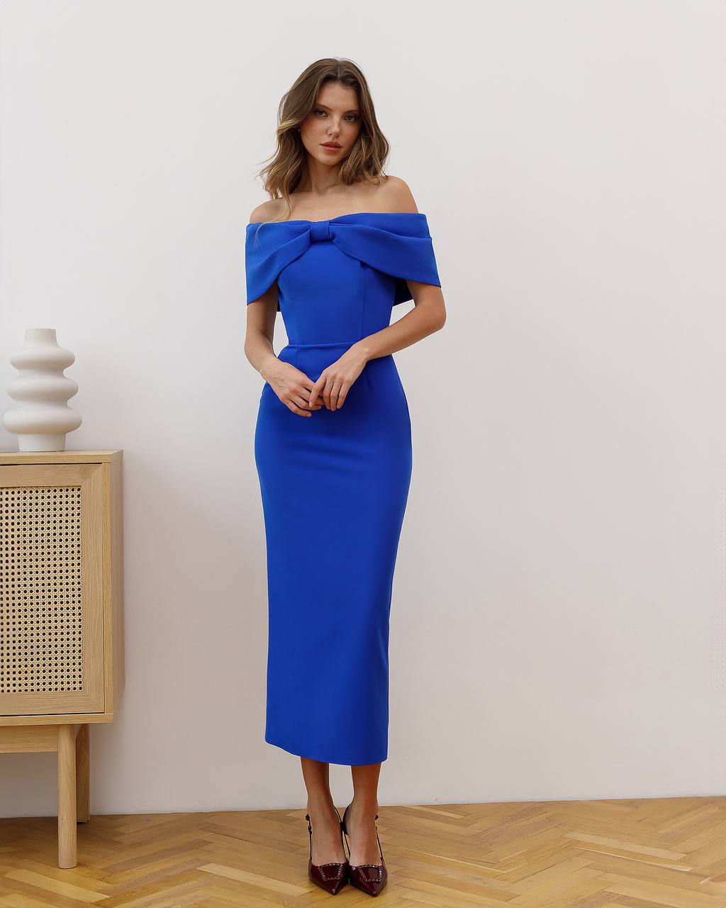 Woman wearing a blue off-shoulder dress in a room with a wooden cabinet and decorative items.