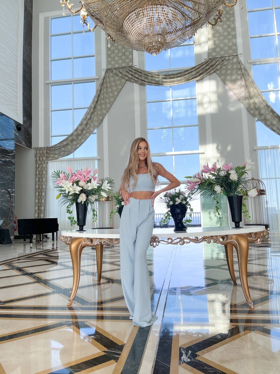 Woman in a stylish outfit standing in a luxurious room with large windows and decorative flowers.