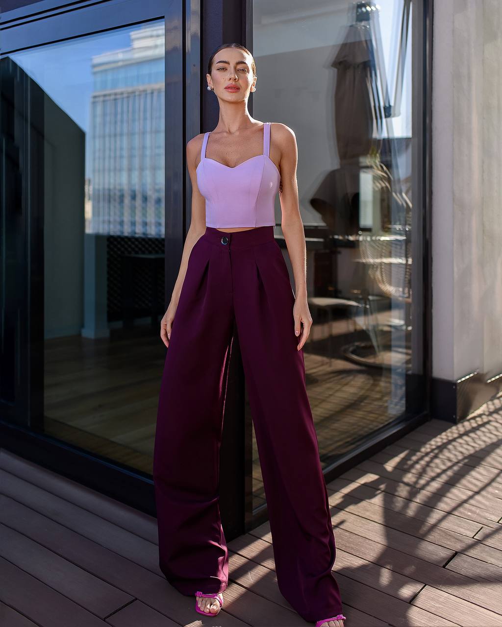 Woman in a light purple top and dark purple pants standing on a wooden deck with glass walls.