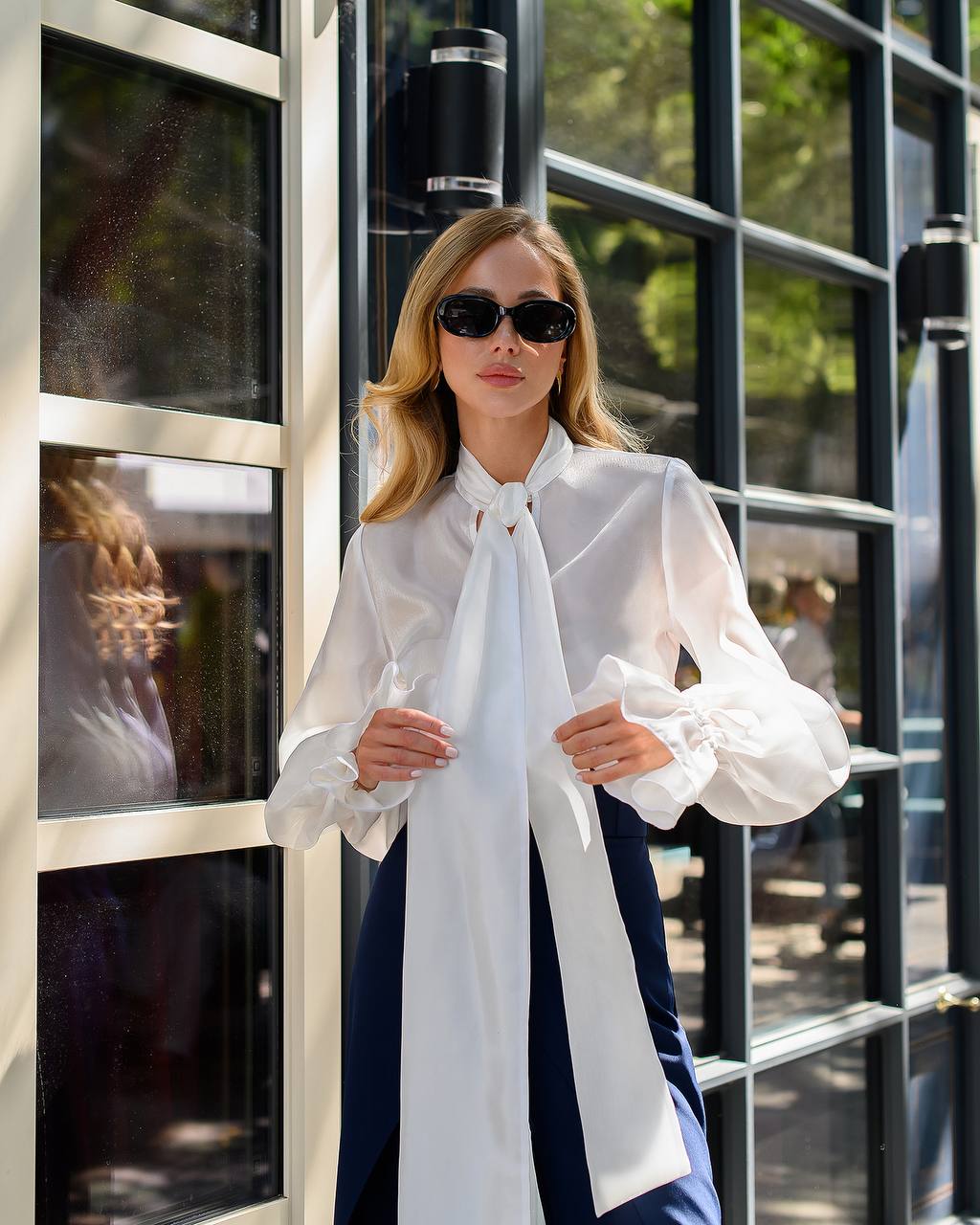 Woman wearing a white blouse with a large bow tie in front of a glass door.
