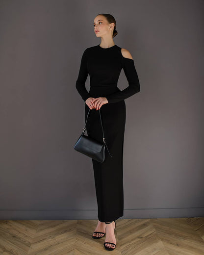 Woman in a black outfit holding a black handbag against a gray wall.