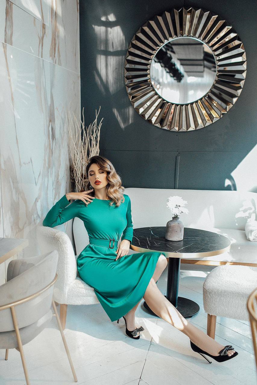 Woman in a green dress sitting in a stylish interior setting with a large mirror on the wall.
