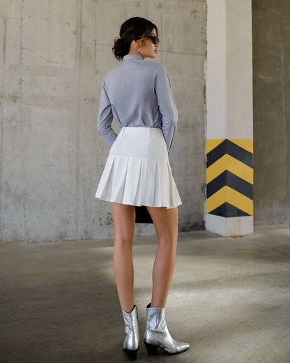 Woman wearing a light blue shirt, white pleated skirt, and silver boots in an industrial setting.