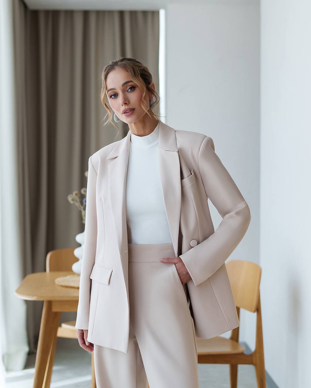 Woman wearing a beige suit standing in a room with a table and chairs.