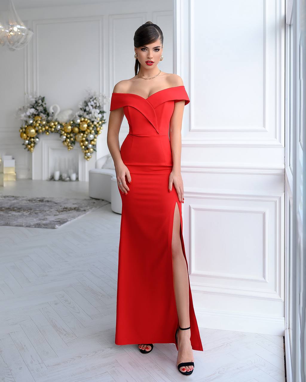 Woman in a red off-shoulder gown with a high slit standing in a stylish room.