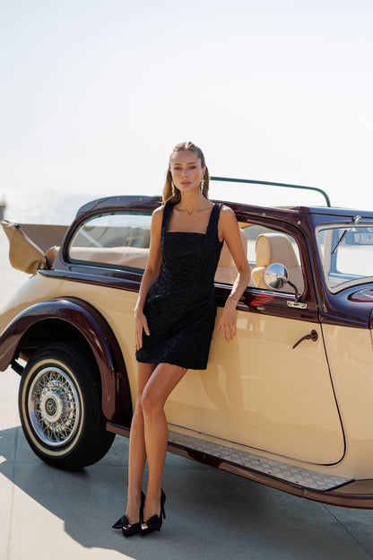 Woman in a black dress standing next to an old car on a sunny day.