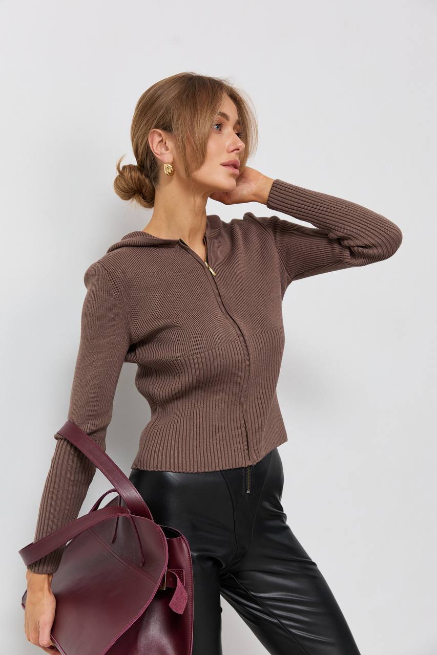 Woman wearing a brown ribbed sweater holding a maroon bag against a white background