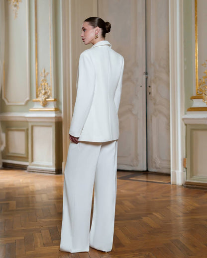 Person wearing a white suit standing in an elegant room with wooden floors and decorative walls.