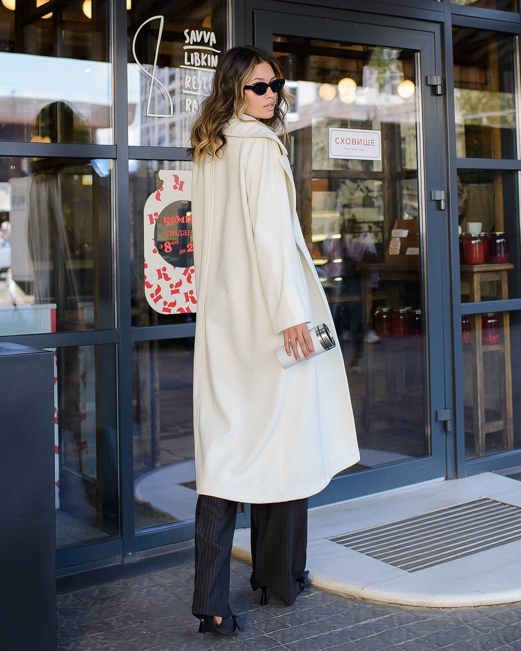 Woman in a white coat standing outside a store with glass doors.