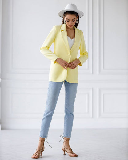 A woman modeling a light yellow blazer, light blue jeans, a white wide-brim hat, and brown strappy heels against a white paneled wall.