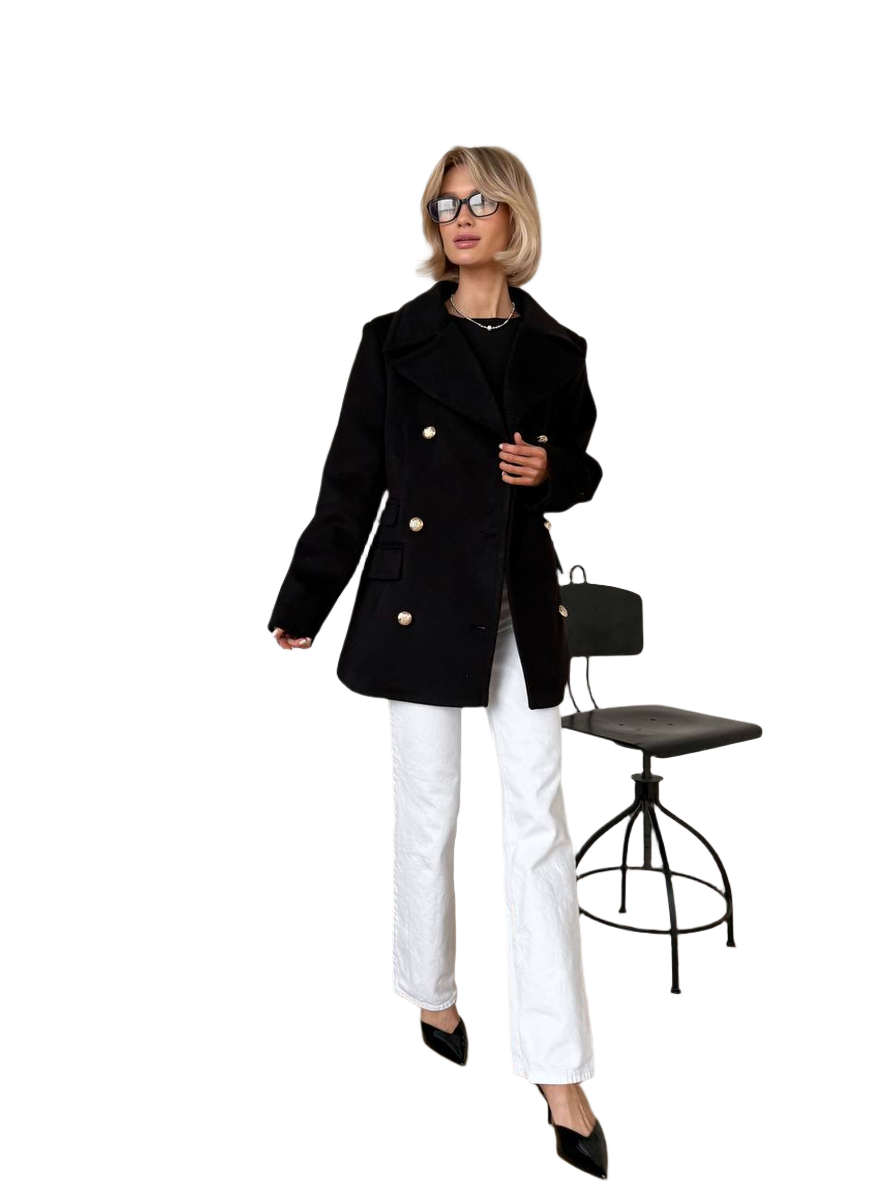 Woman wearing a black coat and white pants standing next to a black chair on a white background
