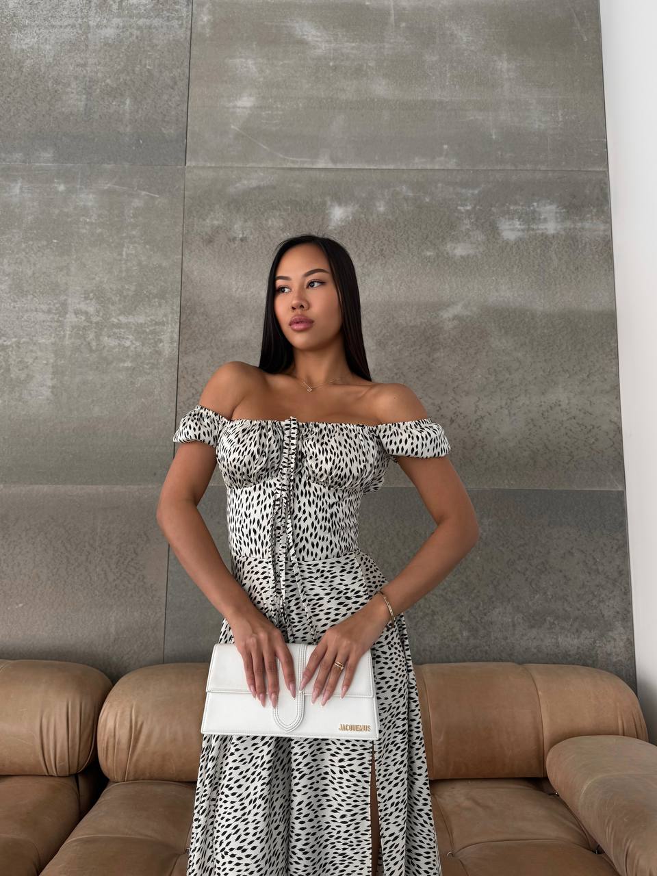 Woman in a patterned dress holding a white clutch against a neutral wall.
