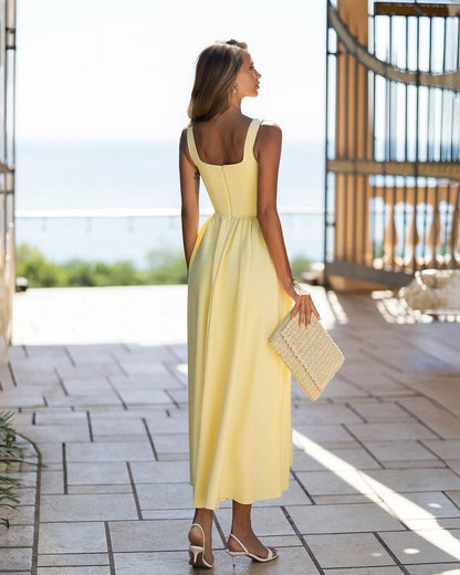 A woman stands on a tiled outdoor terrace wearing a pale yellow sleeveless maxi dress with a scoop neckline and a fitted waist. She is shown from the back, holding a woven clutch and wearing beige high-heeled sandals. The background features a bright, sunlit patio area with a railing overlooking water.