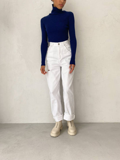 Person wearing a navy turtleneck, white jeans, and white boots against a plain background