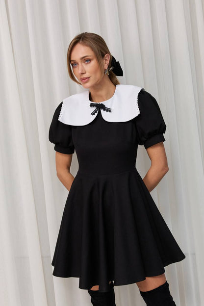 Woman wearing a black dress with a white collar against a white curtain background