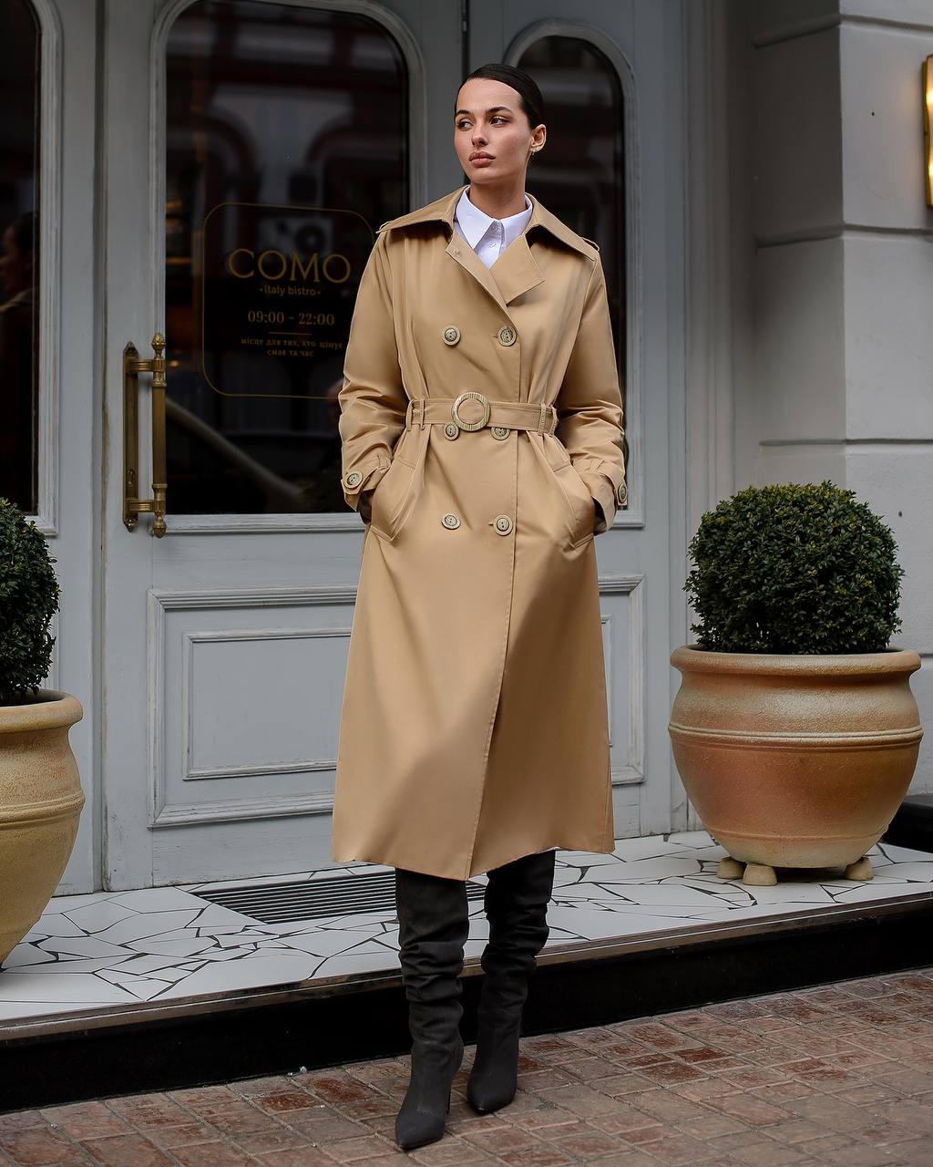 Person wearing a beige trench coat standing in front of a building entrance.