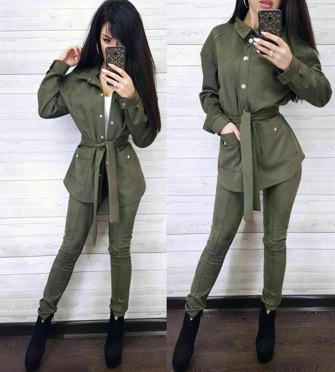 Woman wearing a green outfit with a belt, taking a mirror selfie against a white wooden panel background.
