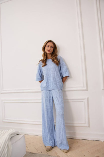 Woman wearing a light blue outfit standing against a white paneled wall.