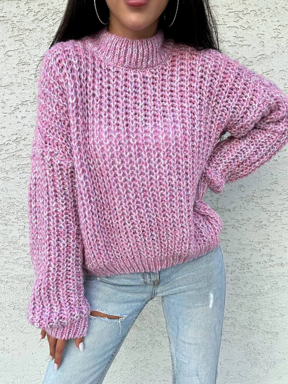 Person wearing a pink knitted sweater and light blue jeans against a textured wall.