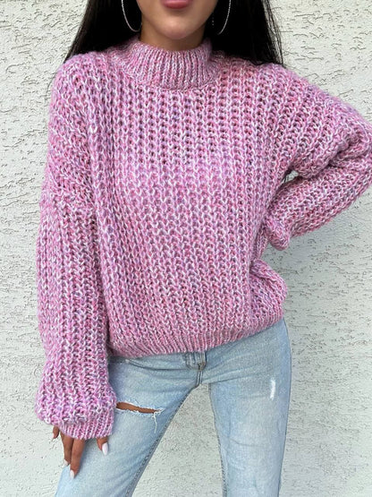 Person wearing a pink knitted sweater and light blue jeans against a textured wall.