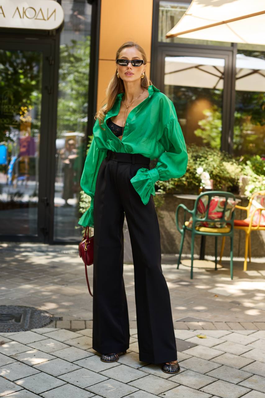 Woman in a green shirt and black pants standing on a street with outdoor seating in the background.