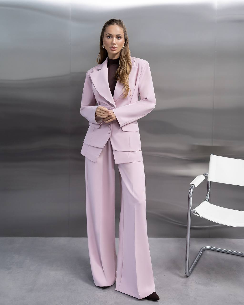 Woman in a pink suit standing in front of a gray background with a white chair.