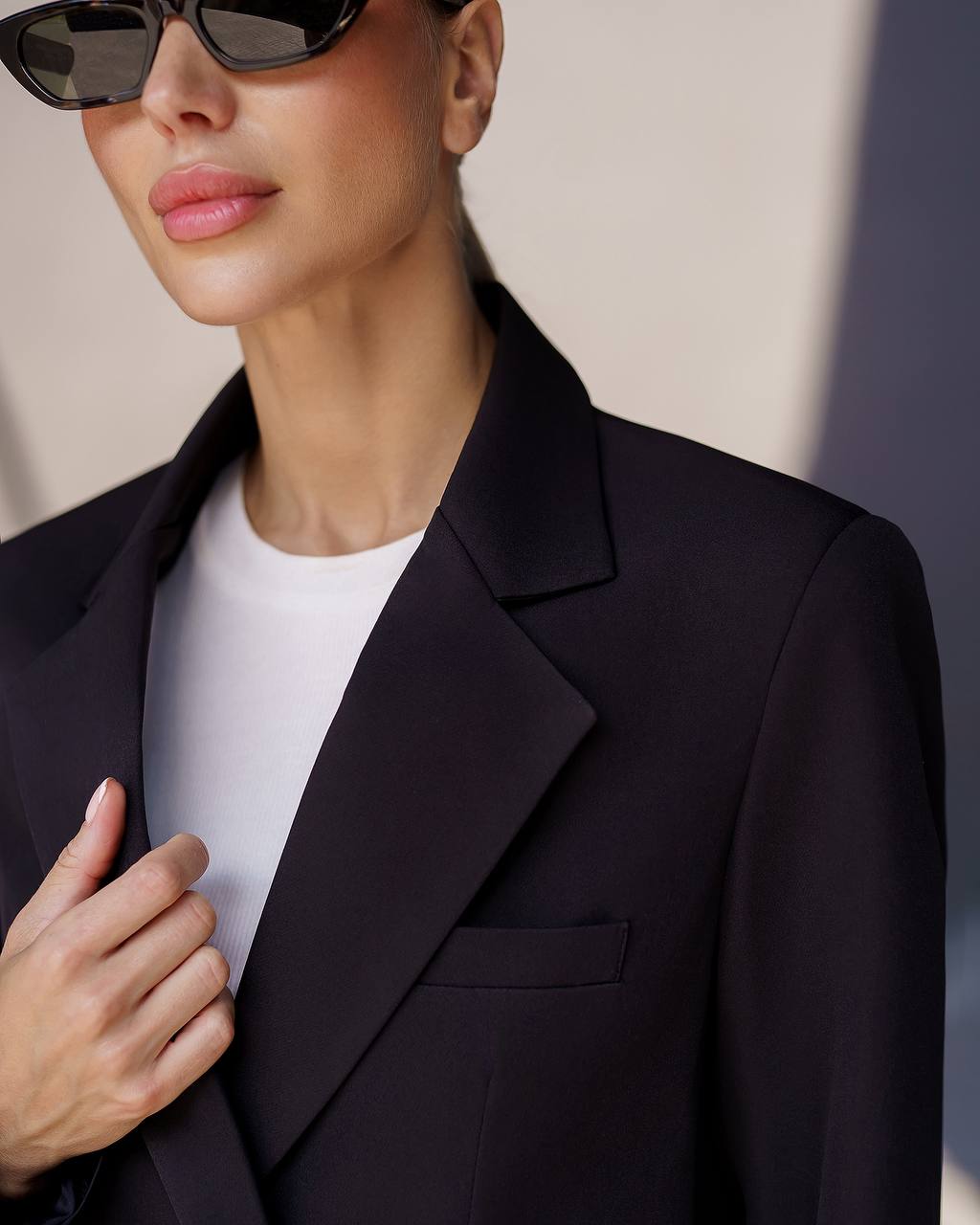 Person wearing a black blazer over a white shirt with sunglasses on a neutral background