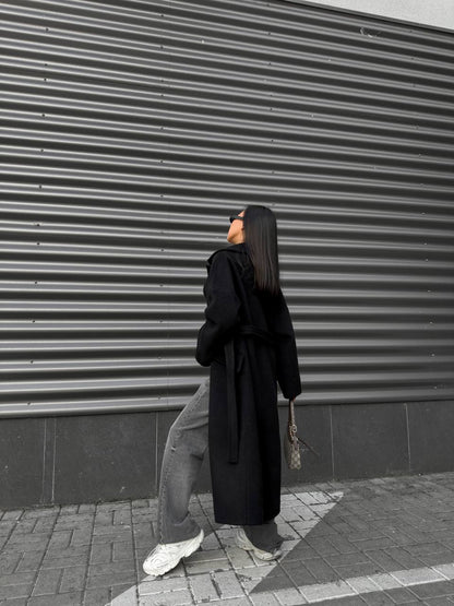 Person wearing a long black coat and light-colored pants standing against a gray corrugated metal wall.