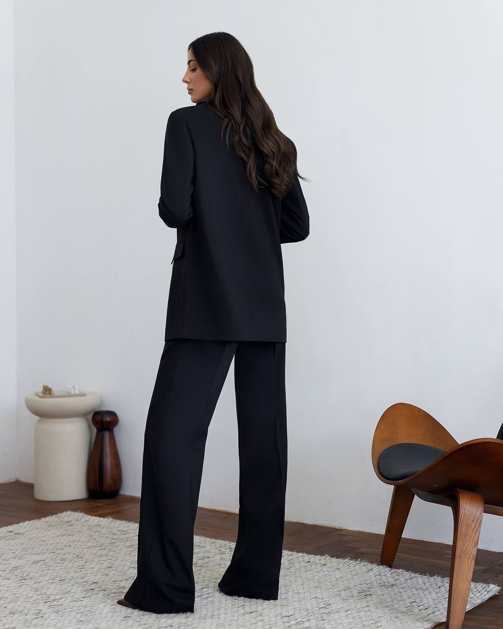 Woman wearing a black suit standing in a room with a wooden chair and decorative items.