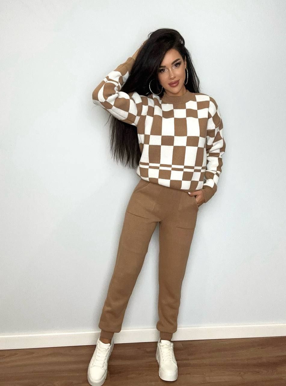 Woman wearing a checkered sweater and brown pants against a white wall.