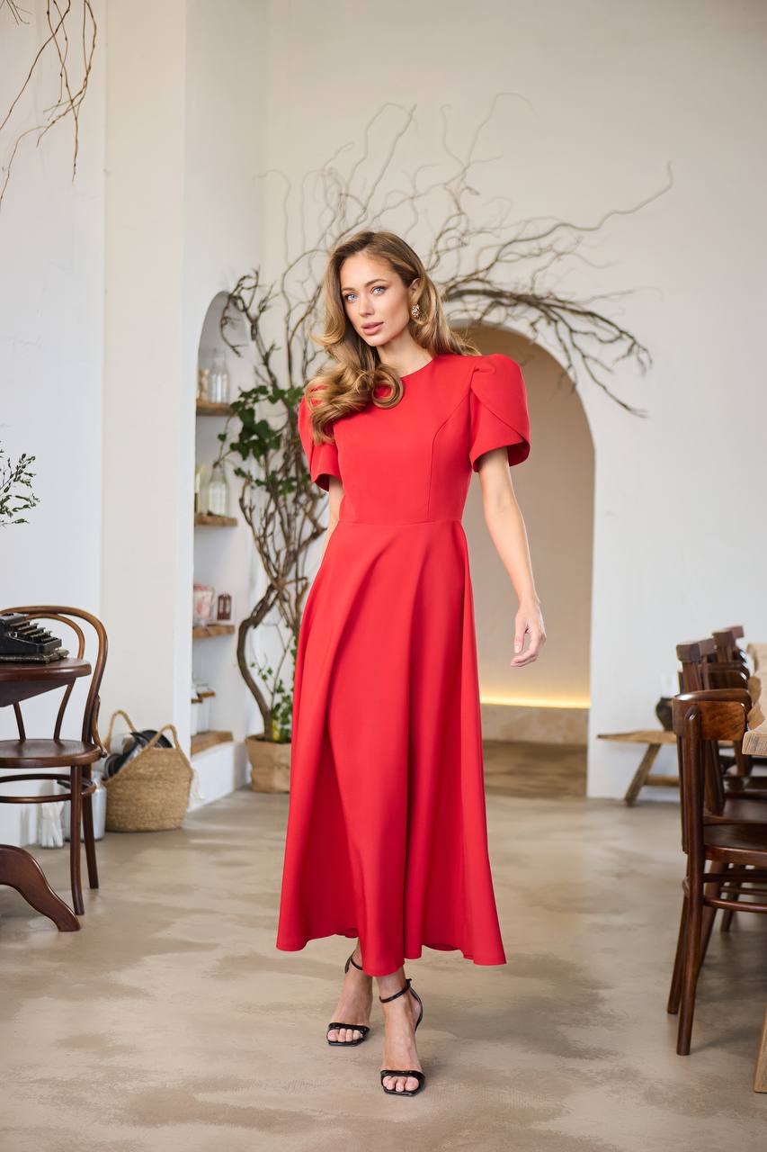 Woman in a red dress standing in a room with wooden furniture and decorative plants.