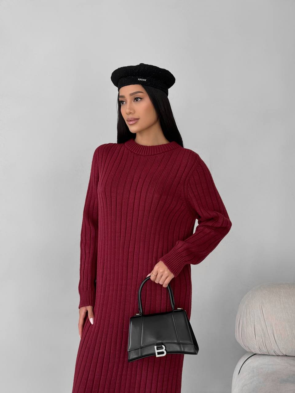 Woman wearing a burgundy ribbed dress and black beret holding a black handbag against a gray background
