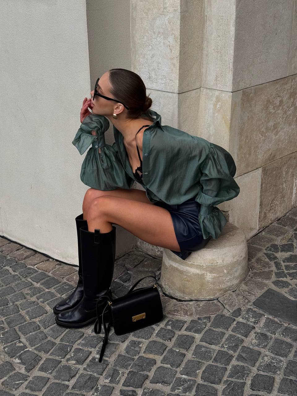 Woman sitting on steps wearing a green outfit with black boots and a black handbag.