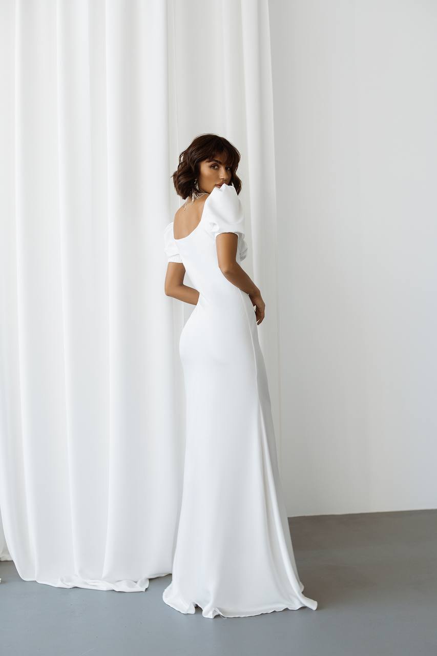 Woman wearing a white off-shoulder gown standing against a white curtain.