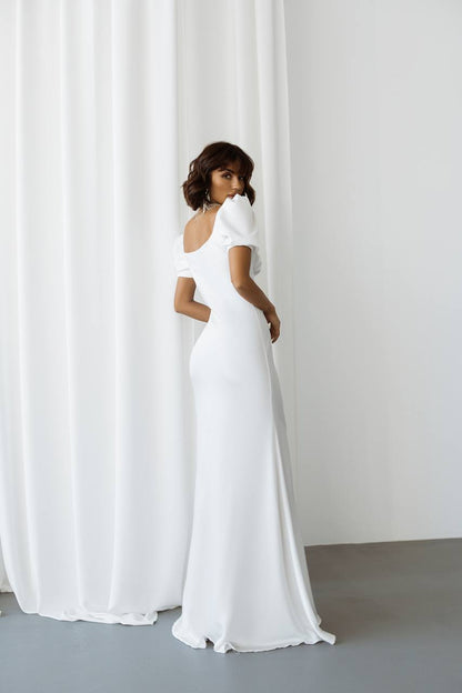 Woman wearing a white off-shoulder gown standing against a white curtain.