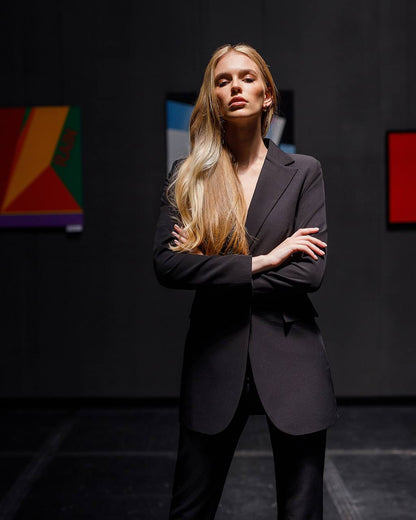 Woman in a black suit standing against a dark background with colorful abstract art.