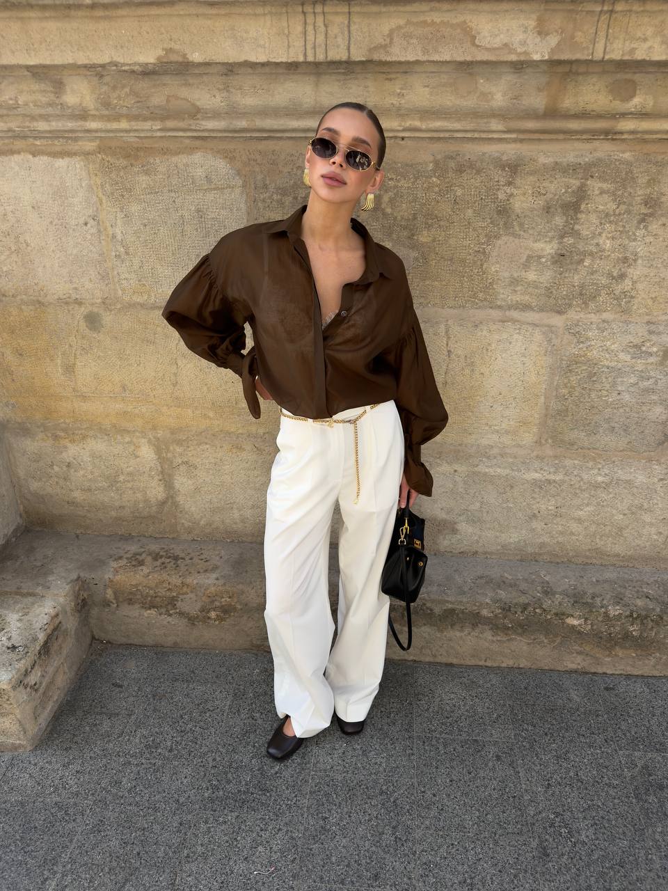 Woman in brown shirt and white pants standing against a stone wall.