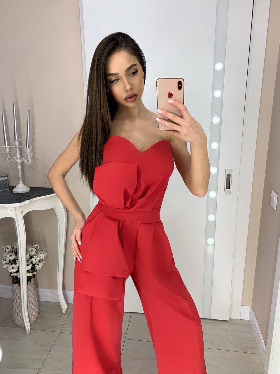 Woman taking a mirror selfie wearing a red strapless jumpsuit in a room with decorative elements.