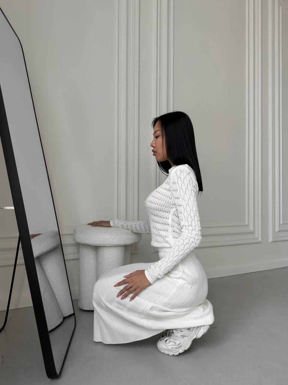 Person in a white outfit posing in a minimalistic room with a white wall and gray floor.