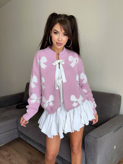 Woman wearing a pink cardigan with white bow patterns and a white skirt indoors.