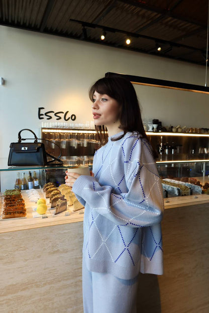 Woman in a cafe wearing a light blue sweater with a patterned design, standing in front of a display case with various items.