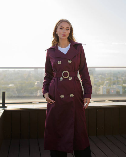 Woman wearing a burgundy trench coat with gold buttons on a rooftop with cityscape view.
