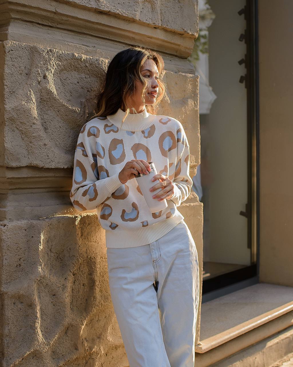 Woman wearing a patterned sweater and white pants standing against a stone wall.